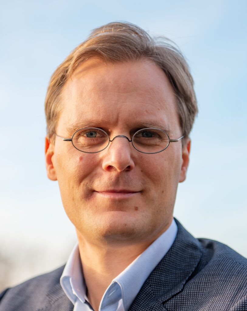 „Porträt von Jonas Niemann. Er blickt in die Kamera und trägt eine Brille, hellblaues Hemd und dunkles Sakko. Der Hintergrund ist unscharf.“